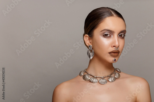 Frontal beauty portrait of a girl with makeup and jewelry applied on face, with bare shoulders, over gray background.