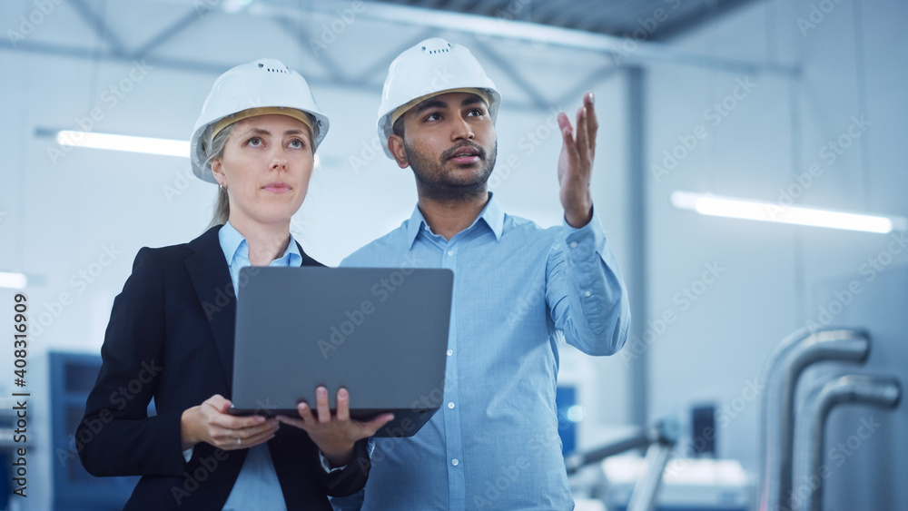 Female Chief Engineer and Male Project Managers Standing in the Middle of Modern Factory, Use Laptop Computer, Plan Production Line Optimization. Professional People work on Industrial CNC Machinery Female Chief Engineer and Male Project Managers Standing in the Middle of Modern Factory, Use Laptop Computer, Plan Production Line Optimization. Professional People work on Industrial CNC Machinery