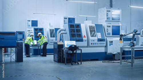 Chief Engineer and Project Manager Wearing Safety Vests and Hard Hats Work on Computer, Talk, Optimize Production Line. On Foreground: industry 4.0 Modern Factory with CNC Machinery and robot arm