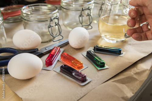 Frau mit Essig, Wasser und bunten Ostereier Kaltfarben in Rot, Orange, Gelb, Blau, Grün in Verpackung auf Papier Unterlage mit Gläser, gekochte Eier und Schere zum Färben zu Ostern