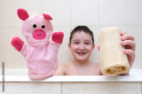 A boy with a funny face sitting in a bathtub and showing loofah and hand puppet washcloth close up photo