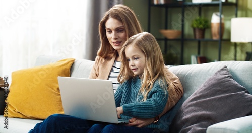 Happy mother and cute little daughter doing interesting tasks together using laptop sitting at home on couch.