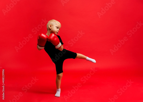 a little boy boxer stands in boxing gloves and makes a lunge with his foot on a red background with a place for text