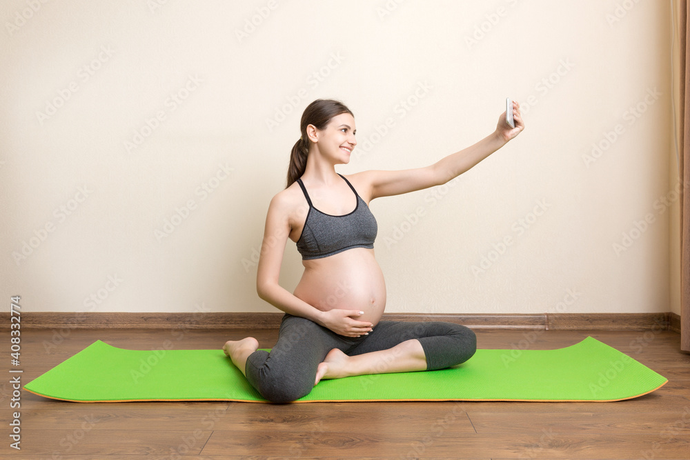 Pregnant female holding smartphone while sitting on exercise yoga mat at home. Fitness or yoga training online at coronavirus time Pregnant female holding smartphone while sitting on exercise yoga mat at home. Fitness or yoga training online at coronavirus time