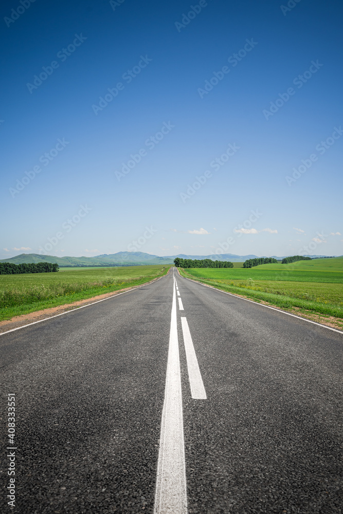 An asphalt road among meadows leading to the mountains on the horizon. Beautiful summer landscape with blue skies, greenery and highway. Minimalism, transport links, travel concept. An asphalt road among meadows leading to the mountains on the horizon. Beautiful summer landscape with blue skies, greenery and highway. Minimalism, transport links, travel concept.