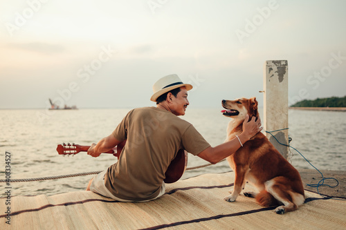 A man wear straw hat and playing guitar music song near the sea sunset and stroking the head dog pet.
