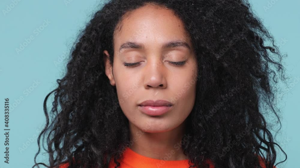 Close up young calm african american woman with curly hair lush hairstyle 20s years old in orange sweatshirt open eyes wake up looking camera isolated on pastel blue color background studio portrait