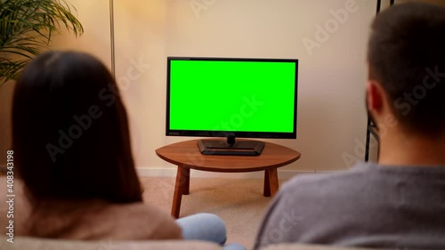 Caucasian Couple Sitting Together On Couch At Home In A Cozy Room Watching Green Chroma Key Screen. Looking TV Show Or News In Home Rest Over The Shoulder Shot