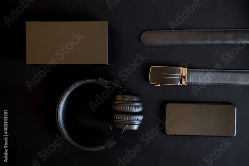 A men's black leather belt with a golden automatic buckle, a smartphone, wireless headphones with ear pads and a gift box lie on a dark background.