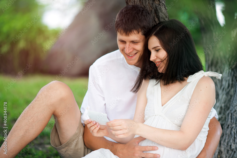 A pregnant girl and a guy sit under a tree in the park and look at the ultrasound photo of the unborn child. A young happy Caucasian family is waiting for the birth of a baby. Husband gently embracing A pregnant girl and a guy sit under a tree in the park and look at the ultrasound photo of the unborn child. A young happy Caucasian family is waiting for the birth of a baby. Husband gently embracing