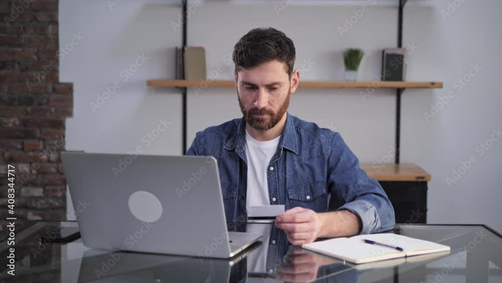 Handsome caucasian bearded man sitting in front of laptop holding his bank credit card doing online shopping purchase buying stuff typing card number on living background. Handsome caucasian bearded man sitting in front of laptop holding his bank credit card doing online shopping purchase buying stuff typing card number on living background.