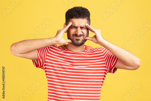 Frustrated unhealthy bearded man in striped t-shirt pressing fingers to temples, feeling sick and suffering severe headache, migraine of stress, tension