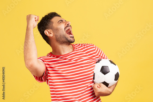 Extremely positive satisfied man in striped red t-shirt screaming holding soccer ball, celebrating victory of favourite football team on championship