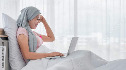 Young Asian woman wearing a hijab uses smartphone and laptop at her desk. Attaching a pink ribbon represents recovery from a breast cancer patient. Breast cancer concept, cancer prevention concept.