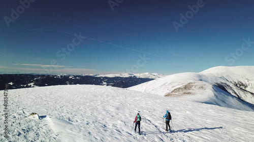 A drone shot of a couple wearing snow shoes hiking up to the peak of Speikkogel in Austrian Alps. The whole slope is covered with snow. many mountain chains in the back. Winter outdoor activity. Fun