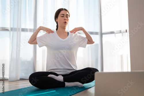 A young woman in sportswear, sitting doing yoga on online lessons. Indoor. Windows on the background. The concept of online training at home