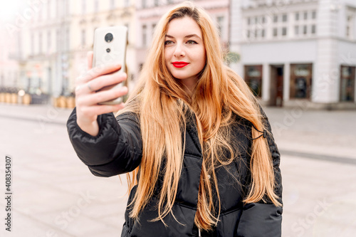 Beautiful girl takes selfie photo. Blonde with phone on city street. Young woman posing for social networks