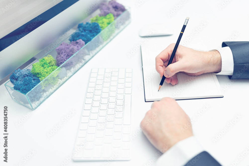 Young man in suit writing in pencil on notebook at computer desk. Business concept Young man in suit writing in pencil on notebook at computer desk. Business concept