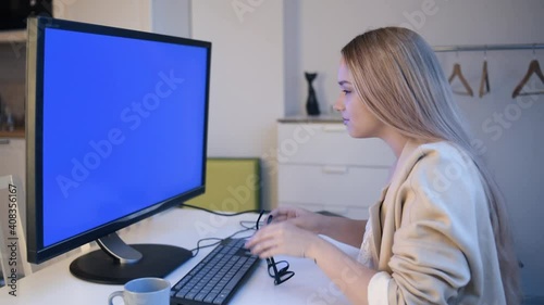 The girl looks into the green screen of the monitor. A beautiful blonde with glasses works at home in front of a large computer. The woman takes off her glasses and massages her eyes. Eye fatigue