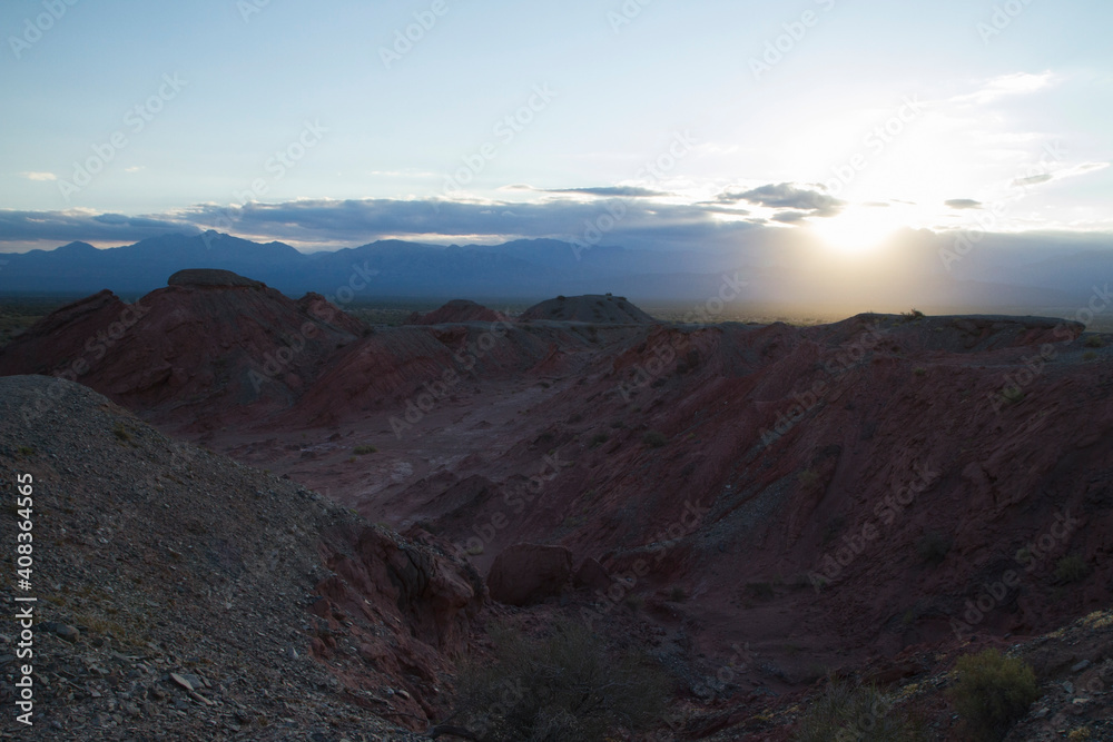 Nightfall in the desert. Beautiful view of the sun hiding in the mountains, the red canyon, dunes and rocky formations at sunset. 