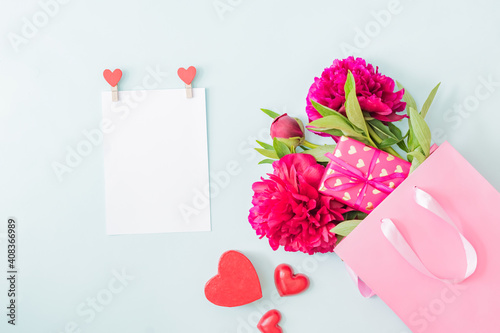 Mockup white greeting card with red peonies in a pink paper bag and gift box on a light background
