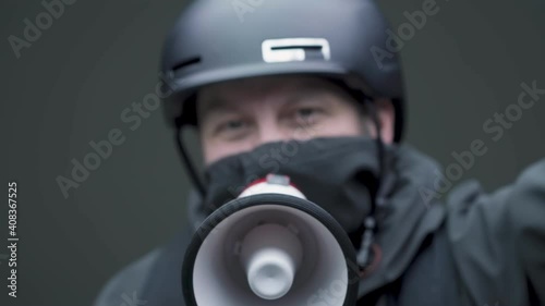 Protesting angry aggressive young caucasian male 30s in black helmet, mask screaming in megaphone, raising up fist photo