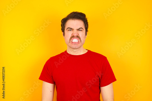 young Caucasian man wearing red t-shirt standing against yellow background keeps teeth clenched, frowns face in dissatisfaction, irritated because of much duties.