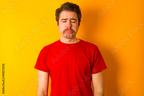 Dismal gloomy rejected young Caucasian man wearing red t-shirt standing against yellow background has problems and difficulties, curves lower lip and closes eyes in despair, being in depression