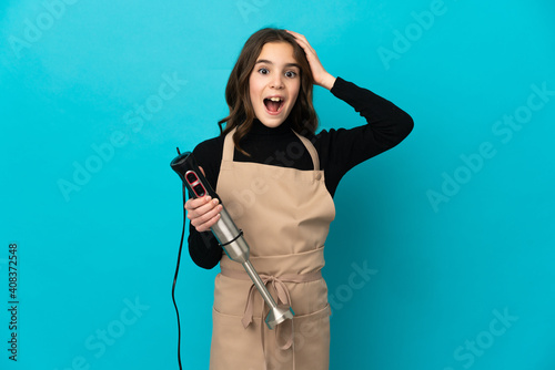 Little girl using hand blender isolated on blue background with surprise expression