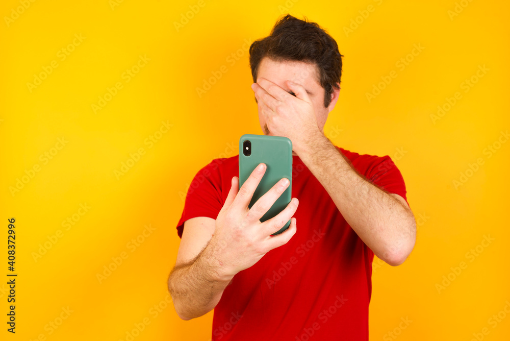 Young Caucasian man wearing red t-shirt standing against yellow background looking at smart phone feeling sad holding hand on face. Young Caucasian man wearing red t-shirt standing against yellow background looking at smart phone feeling sad holding hand on face.