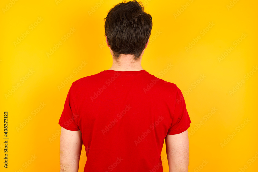 The back view of a Young Caucasian man wearing red t-shirt standing against yellow background. Studio Shoot. The back view of a Young Caucasian man wearing red t-shirt standing against yellow background. Studio Shoot.