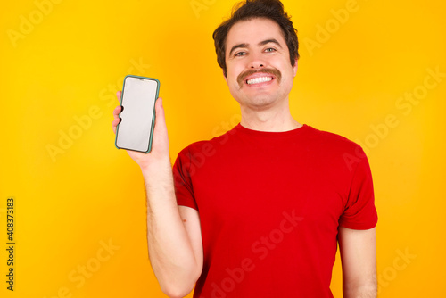 Smiling Young Caucasian man wearing red t-shirt standing against yellow background Mock up copy space. Hold mobile phone with blank empty screen