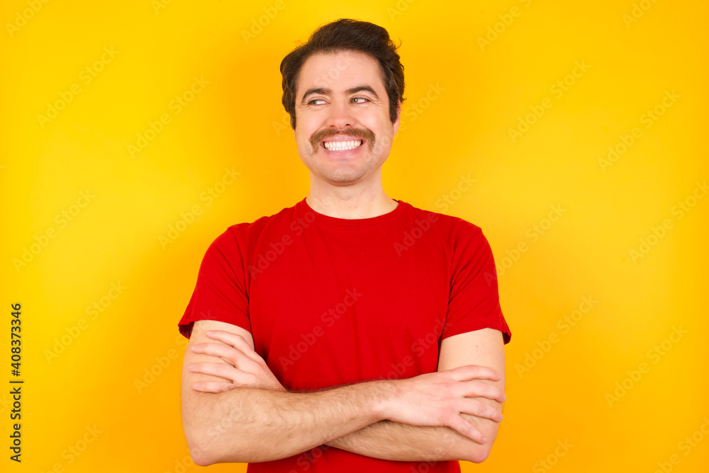 Dreamy rest relaxed Young Caucasian man wearing red t-shirt standing against yellow background crossing arms, looks good copyspace