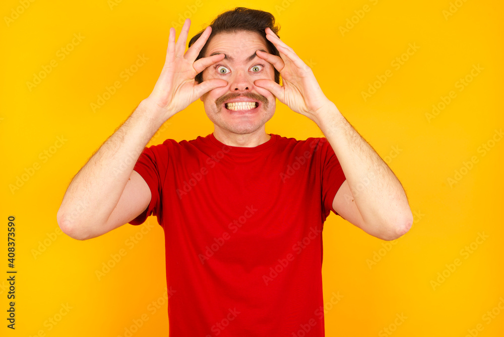 Young Caucasian man wearing red t-shirt standing against yellow background keeping eyes opened to find a success opportunity. Young Caucasian man wearing red t-shirt standing against yellow background keeping eyes opened to find a success opportunity.