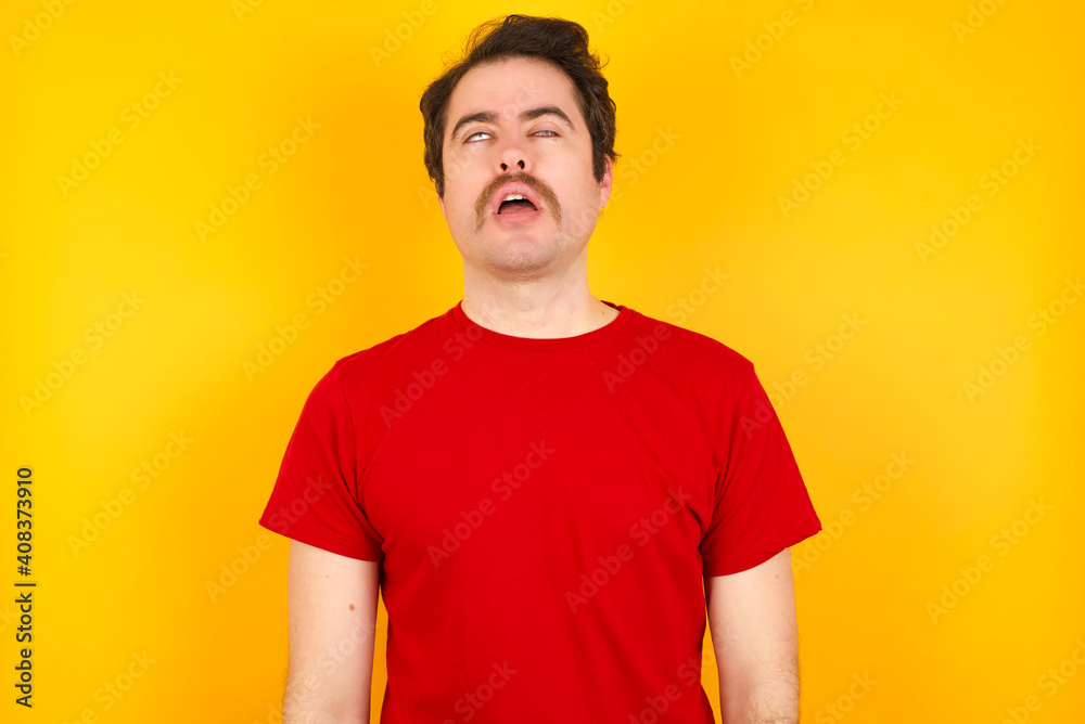 Young Caucasian man wearing red t-shirt standing against yellow background looking sleepy and tired, exhausted for fatigue and hangover, lazy eyes in the morning. Young Caucasian man wearing red t-shirt standing against yellow background looking sleepy and tired, exhausted for fatigue and hangover, lazy eyes in the morning.