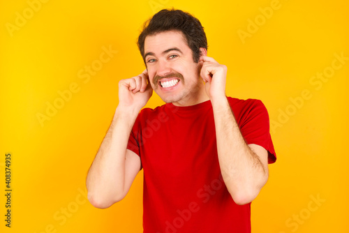 Young Caucasian man wearing red t-shirt standing against yellow wall covering ears with fingers with annoyed expression for the noise of loud music. Deaf concept. © Roquillo