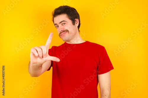 Young Caucasian man wearing red t-shirt standing against yellow wall making fun of people with fingers on forehead doing loser gesture mocking and insulting.