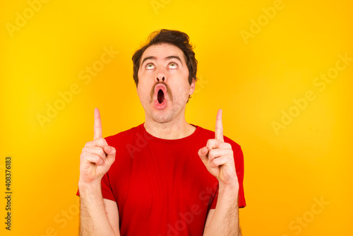 Young Caucasian man wearing red t-shirt standing against yellow wall amazed and surprised looking up and pointing with fingers and raised arms.