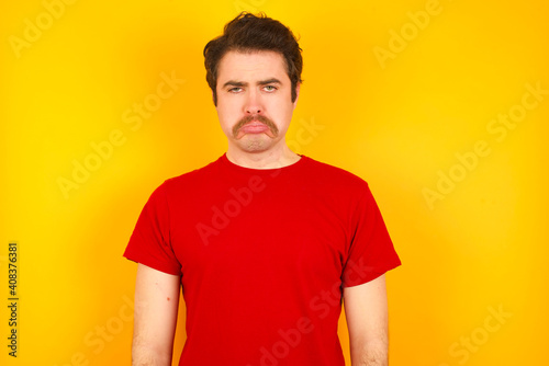 Displeased upset Young Caucasian man wearing red t-shirt standing against yellow wall frowns face as going to cry, being discontent and unhappy as can't achieve goals, Disappointed model has troubles