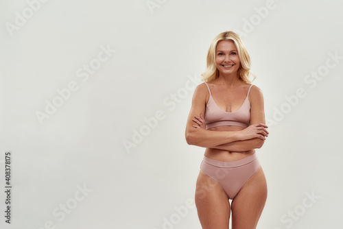 Cheerful beautiful middle aged woman wearing lingerie keeping arms crossed, smiling at camera while posing isolated over grey background © Svitlana