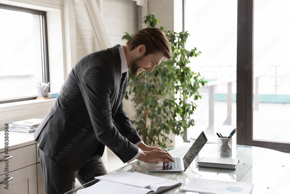 Successful businessman employee worker wearing suit standing at desk, working on laptop in modern office, confident executive manager searching information in internet, writing business email Successful businessman employee worker wearing suit standing at desk, working on laptop in modern office, confident executive manager searching information in internet, writing business email