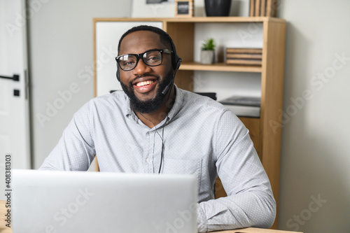 Friendly and handsome African-American wearing glasses and headset in the office, sitting at the desk with an open laptop, working in the customer service department as a call center operator