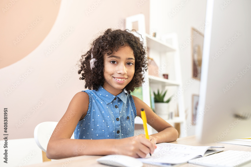 E-learning concept. Cute little multiracial girl looks at the camera and smiles, watching video classes, writing in exercise book, training for an exam with an online coach, studying on the distance E-learning concept. Cute little multiracial girl looks at the camera and smiles, watching video classes, writing in exercise book, training for an exam with an online coach, studying on the distance