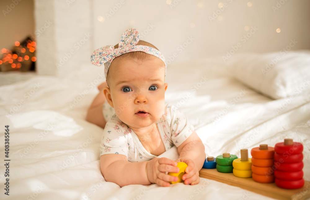 a beautiful baby girl in a colored bodysuit and a headband lies on the bed with an educational toy on her head and looks at the camera a beautiful baby girl in a colored bodysuit and a headband lies on the bed with an educational toy on her head and looks at the camera