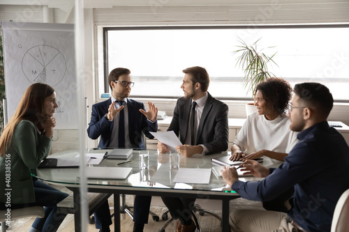 Diverse employees listening to colleague at meeting, sitting at table in boardroom, confident businessman sharing ideas with coworkers at briefing, business partners discussing project strategy