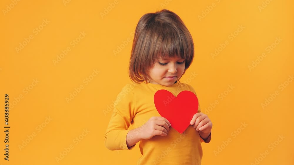 A child with a red heart stands on a yellow background. Little cute boy holds a greeting card in his hand A child with a red heart stands on a yellow background. Little cute boy holds a greeting card in his hand