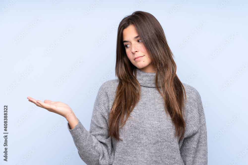 Teenager Brazilian girl over isolated blue background holding copyspace with doubts Teenager Brazilian girl over isolated blue background holding copyspace with doubts