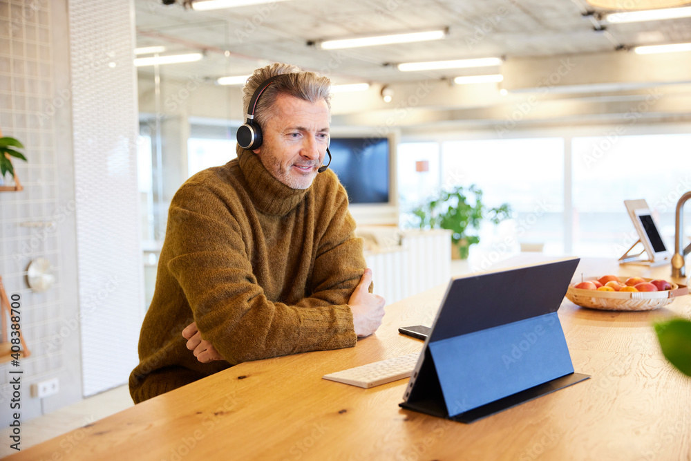 Mature businessman wearing headset working on digital tablet while sitting at office Mature businessman wearing headset working on digital tablet while sitting at office