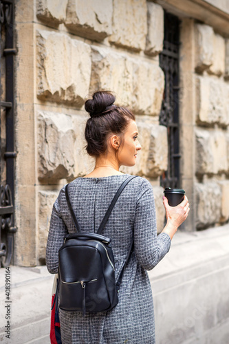 Back view of a young woman with a hot drink and backpack walks in a European city