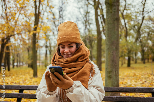 Smiling young woman using mobile phone while sitting on bench at autumn park photo